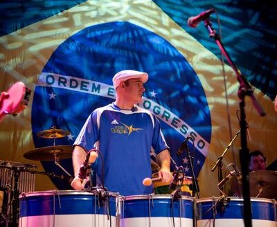 Keynote speaker Josh Robinson performs energetically on Brazilian drums in front of a large Brazilian flag backdrop. Wearing a blue jersey and white cap, Josh leads an engaging musical performance.