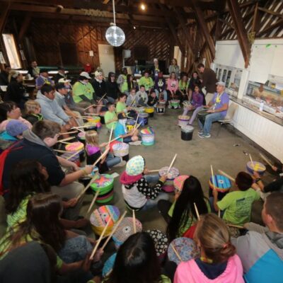 Josh Robinson leads a large group of children and adults in an interactive drumming circle. Participants, seated in a large wooden room, play colorful drums as Josh guides them through the activity, fostering a sense of rhythm, teamwork, and community engagement.