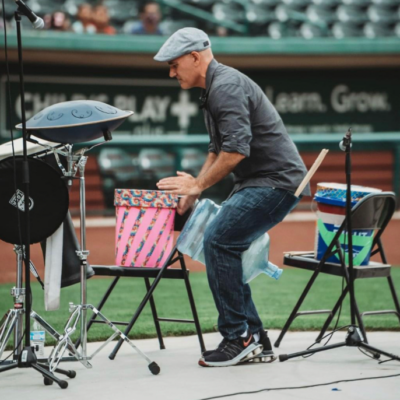 Josh Robinson performs a dynamic drumming session at an outdoor venue, playing various percussion instruments. Wearing a casual outfit and cap, he engages with the drums set up on chairs, delivering an energetic and interactive performance for the audience.