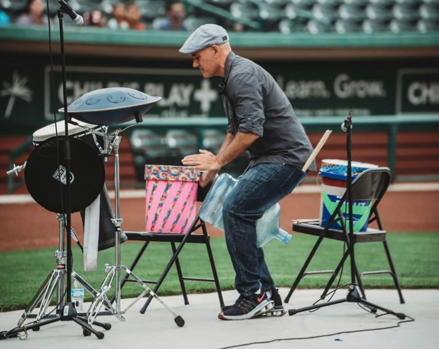 Josh Robinson performs a dynamic drumming session at an outdoor venue, playing various percussion instruments. Wearing a casual outfit and cap, he engages with the drums set up on chairs, delivering an energetic and interactive performance for the audience.