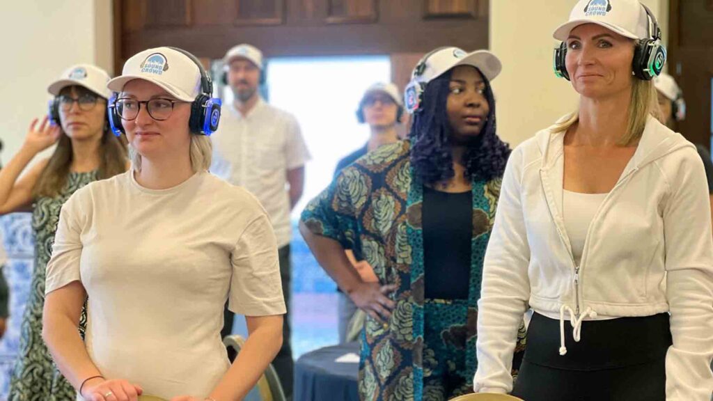 A group of participants in a Sound Crowd team building event stand attentively while wearing noise-cancelling headphones. The activity encourages non-verbal interaction, using sound and movement to foster connection, teamwork, and enthusiasm in a unique way.