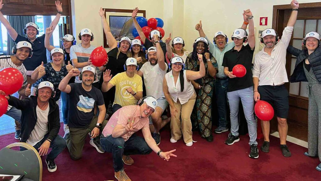 A cheerful group of Sound Crowd participants wearing white caps and headphones pose with balloons and enthusiastic gestures, celebrating the successful conclusion of their team building event. The atmosphere is energetic, and everyone looks engaged and happy.