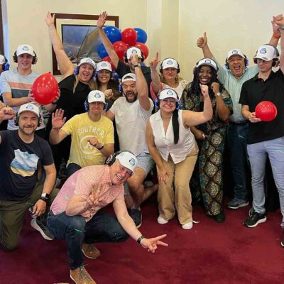 A cheerful group of Sound Crowd participants wearing white caps and headphones pose with balloons and enthusiastic gestures, celebrating the successful conclusion of their team building event. The atmosphere is energetic, and everyone looks engaged and happy.