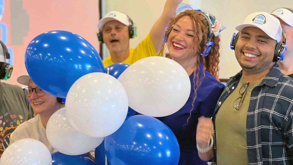 A lively group of Sound Crowd participants smiling and holding blue and white balloons while wearing white caps and headphones during a team building event.