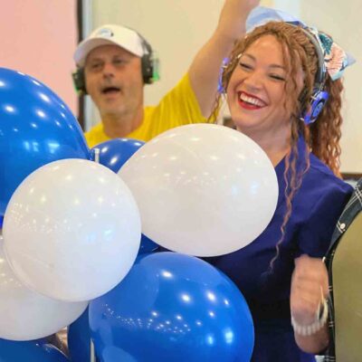 A lively group of Sound Crowd participants smiling and holding blue and white balloons while wearing white caps and headphones during a team building event.