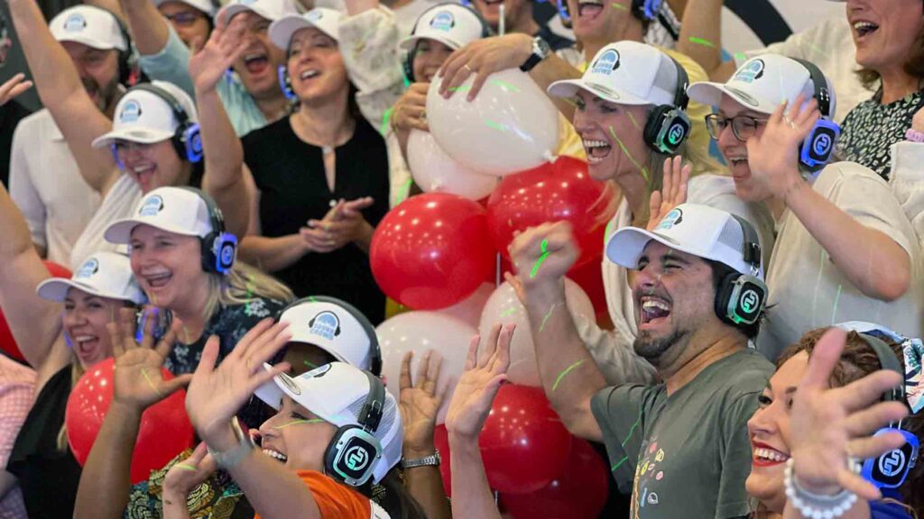 A large group of Sound Crowd participants cheering and smiling while wearing white caps and headphones. They are enthusiastically raising their hands, and red and white balloons are visible in the background, adding to the festive and vibrant atmosphere of the team building event.