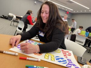 A participant joyfully colors a holiday-themed sign as part of the Hope for the Holidays team building event, creating personalized gift baskets for seniors in local nursing homes.