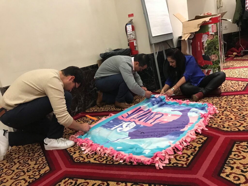 Team members collaborate during the Hope for the Holidays event, carefully crafting a fleece blanket to donate to local seniors as part of their charitable efforts.