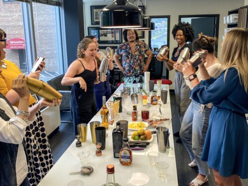 Team shaking cocktail shakers in a mixology class, enjoying the process of creating drinks together. The event promotes collaboration, creativity, and teamwork in a fun and interactive setting.